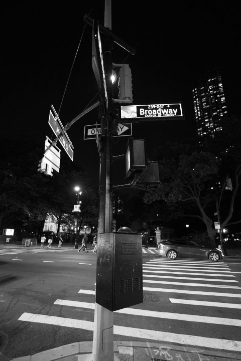 Black and white picture of an intersection sign, at night. The sky is black, a few buildings can be seen, a car driving by, and people walking on the sidewalk.